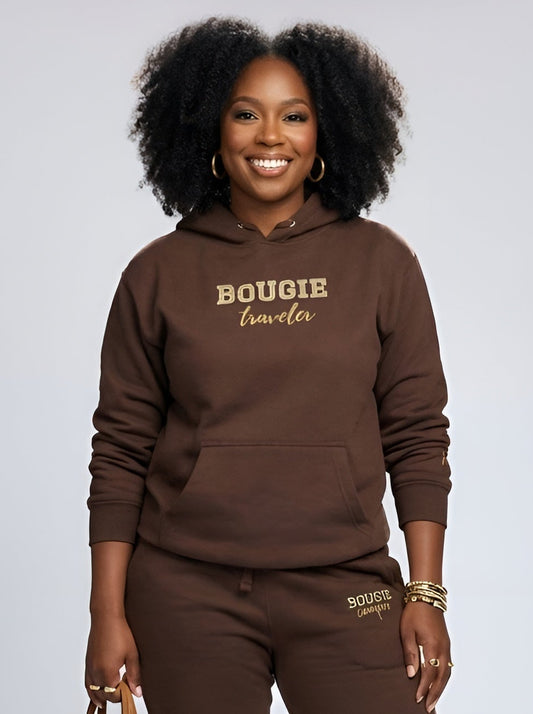 Woman wearing a brown 'Bougie Traveler' hoodie and pants, holding a brown tote bag on a white background.